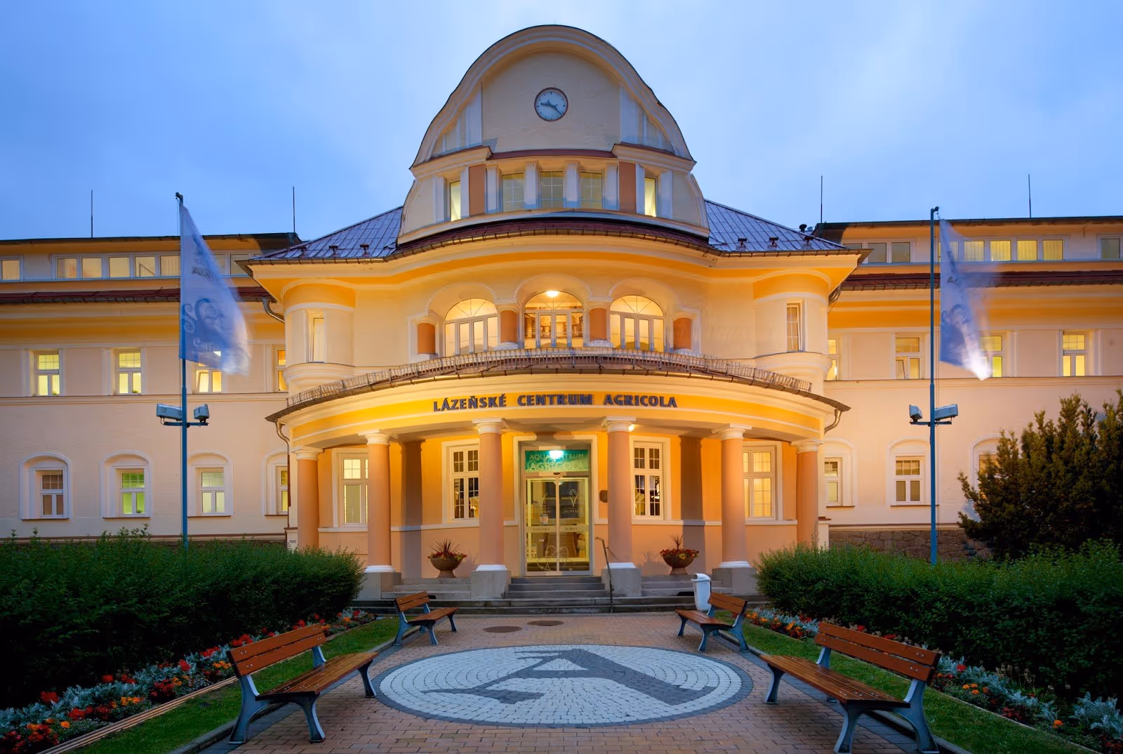 The exterior of Lazeanske centrum Agricola building at dusk with warm lights, pink columns, benches, and two flagpoles in Jachymov Czechia.