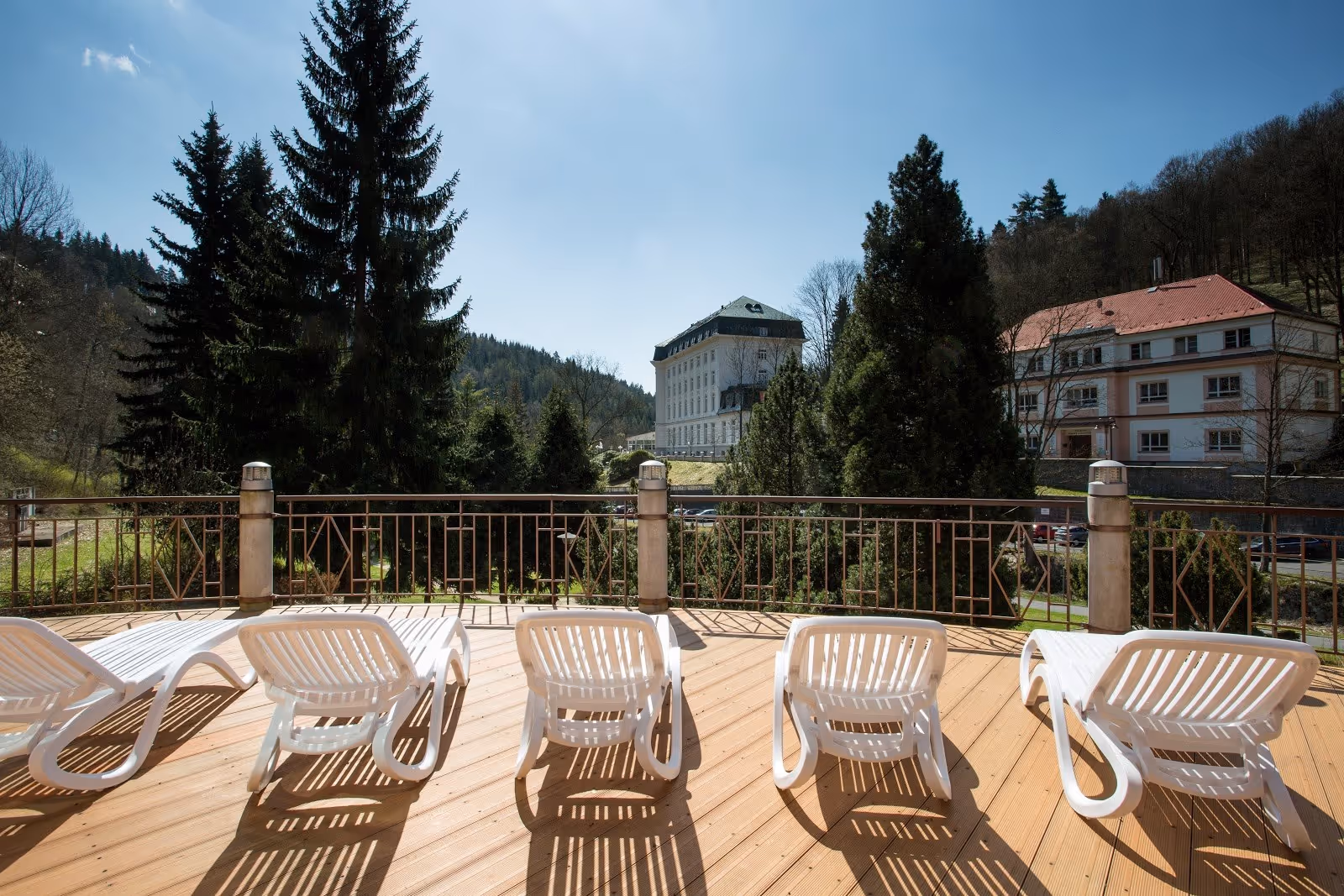 Outdoor lounge area with white plastic chairs on a wooden deck facing tall pine trees and hills near the hot spring center in Jachymov.