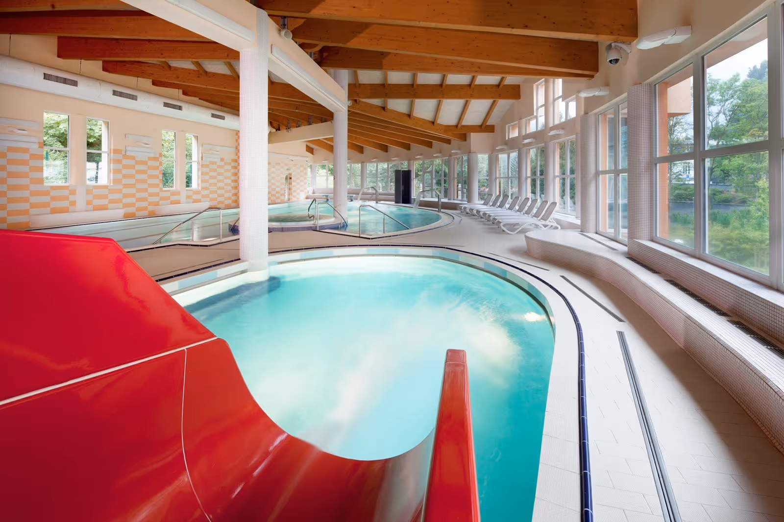 Indoor hot spring pool with turquoise water, curved design, wooden ceiling beams, and large windows overlooking greenery in Jachymov Czechi.