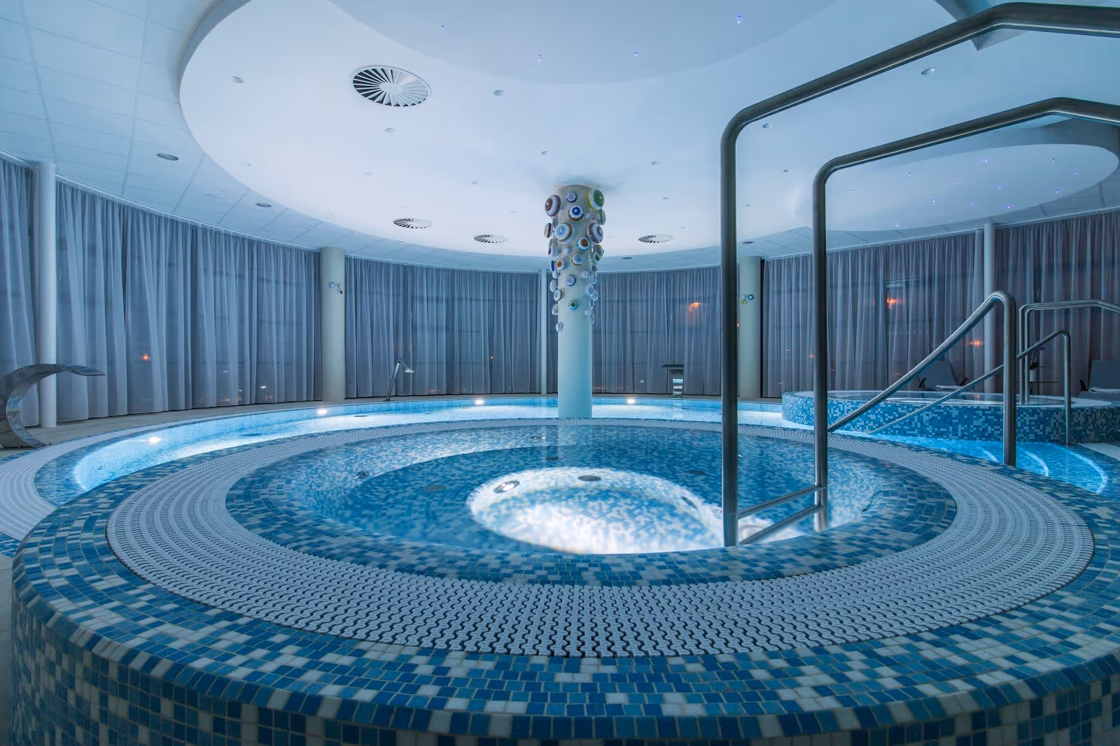 Indoor circular hot spring pool at Lazne Hodonin spa in Hodonin, Czechia, featuring mosaic tiles and metal handrails.