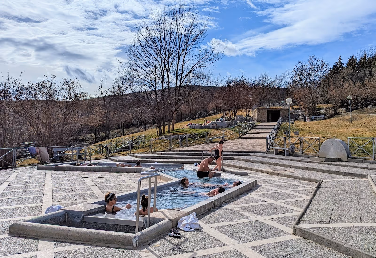 Visitors soaking in a paved Bains de Dorres hot spring pool with leafless trees and natural hillside visible on a cool day in France.
