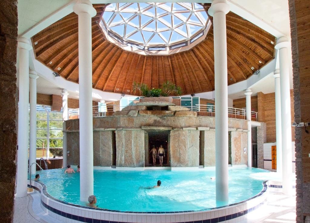 Indoor hot spring pool at Balnea featuring circular stone structure under a wooden dome ceiling with large skylight and white pillars.