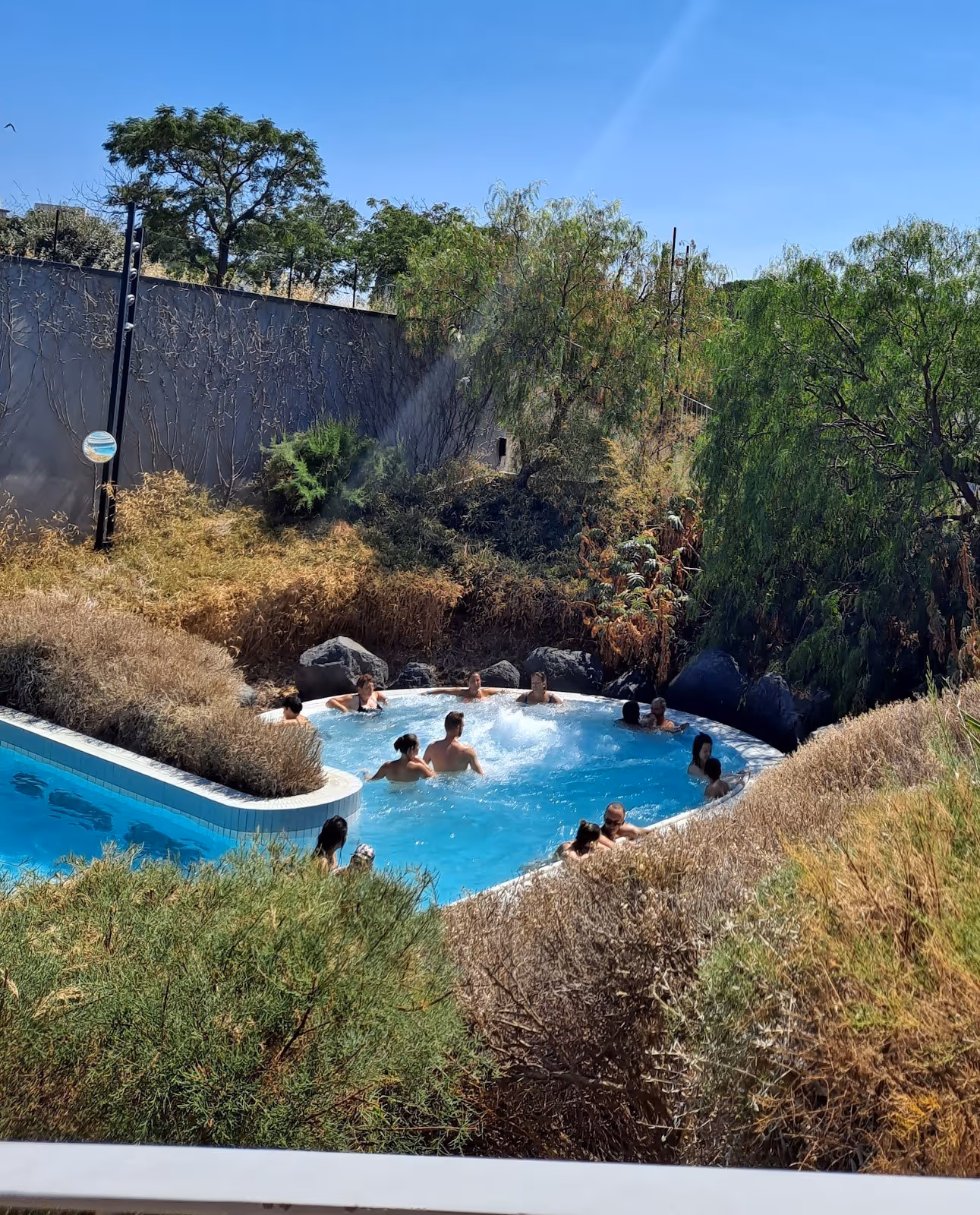 Naturalistic outdoor pool at La Parenthese Obalia surrounded by green trees and shrubs with people enjoying the warm water.