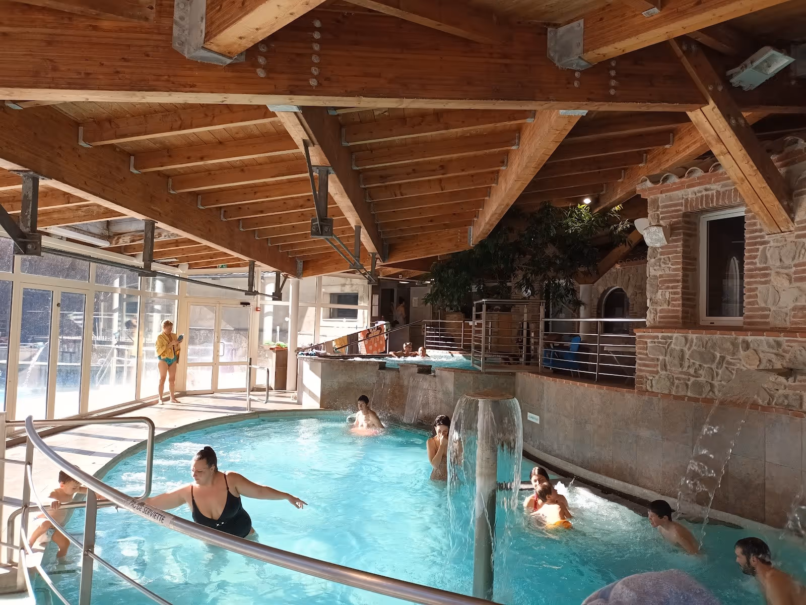 Indoor hot spring pool with wooden beam ceiling at Les Bains de Llo showing people enjoying the water and a wall of large windows letting i.
