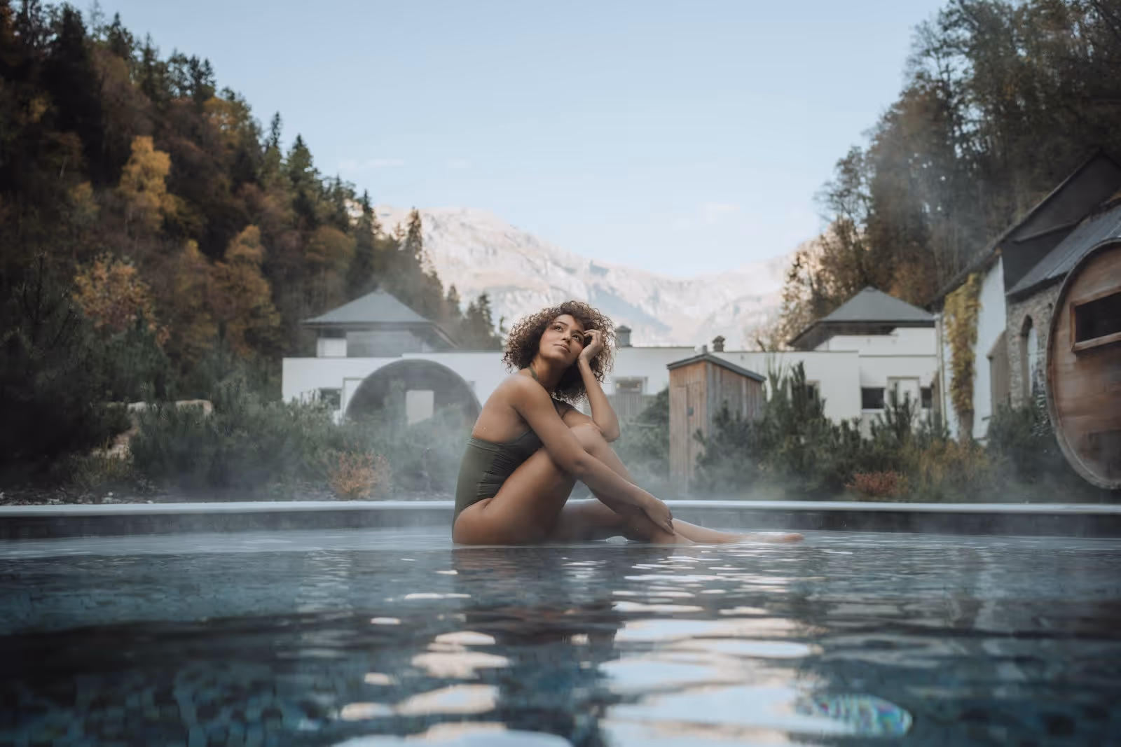 A woman in a dark swimsuit sits relaxed in the steaming outdoor mineral pool at Les Bains du Mont-Blanc near Saint-Gervais-les-Bains, Franc.