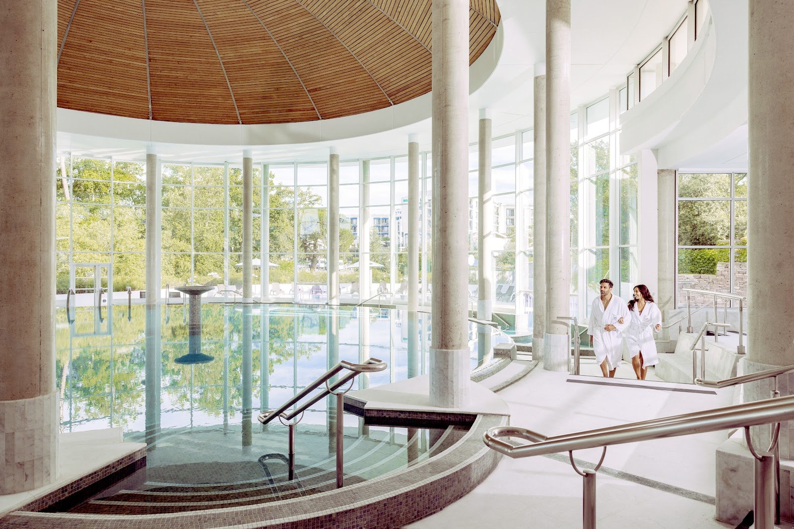Indoor hot spring pool at Sourceo in Dax France with tall glass walls and wooden ceiling showing greenery outside.