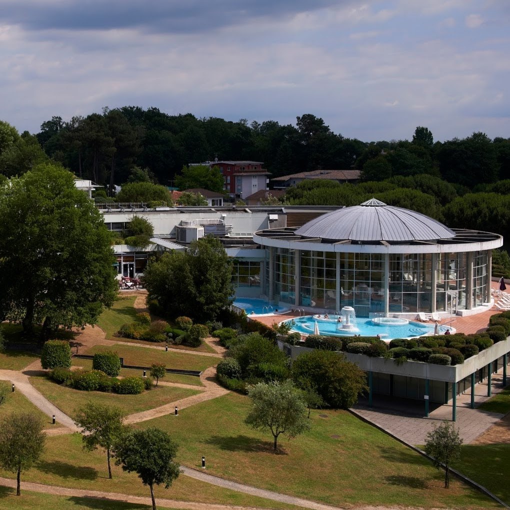 Aerial view of Sourceo hot spring complex in Dax France showing circular glass building with outdoor pools and garden paths.