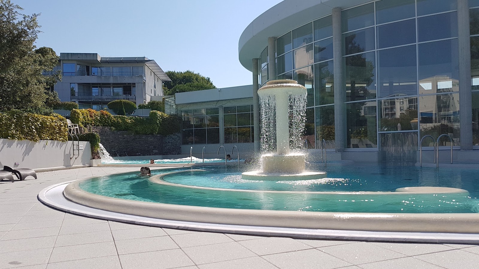 Outdoor spa pool at Sourceo France with round fountain spraying water under bright sunlight near large glass building.