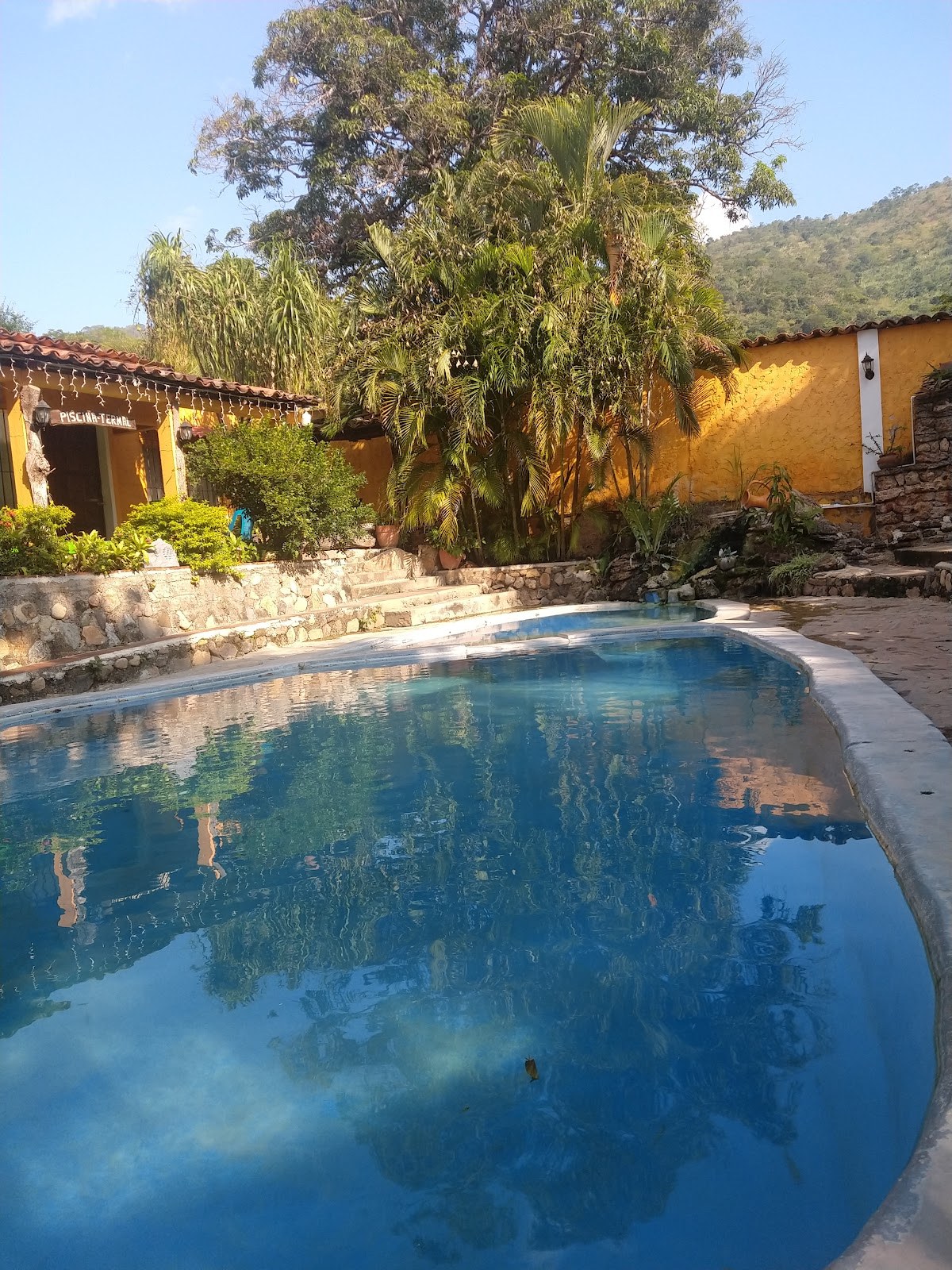 Blue rectangular mineral swimming pool surrounded by stone walls, tropical plants, and yellow buildings at Banos Termales El Brasilar.