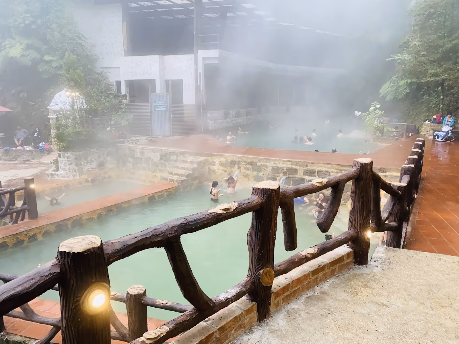 Multiple tiered soaking pools and a wooden fence along a tile walkway with steam rising in the rich green jungle at Las Fuentes Georginas G.