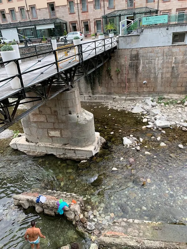 Person soaking in a natural rock lined hot spring pool in a river under a bridge at termas de la hermida in spain