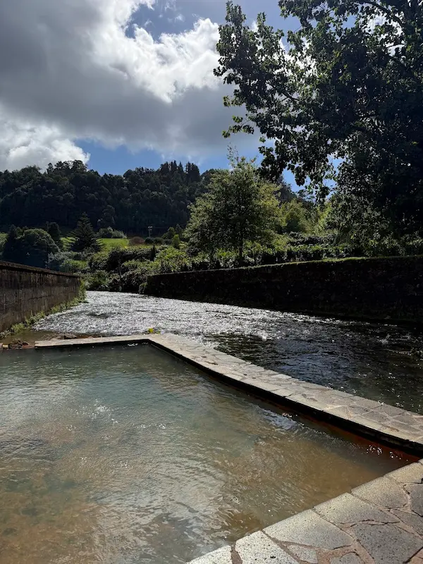 Outdoor concrete hot spring pool at the side of a river with trees in the background at poca da tia silvina in portugal