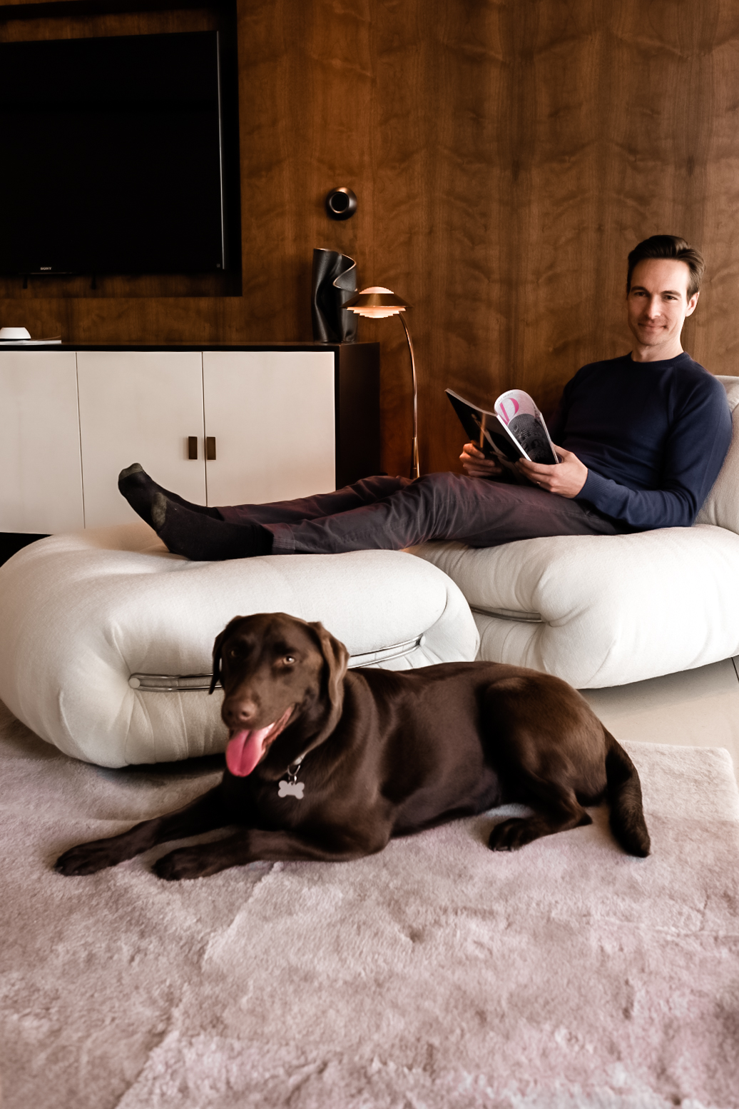 Man reading on a sofa with a dog, wall-mounted television behind