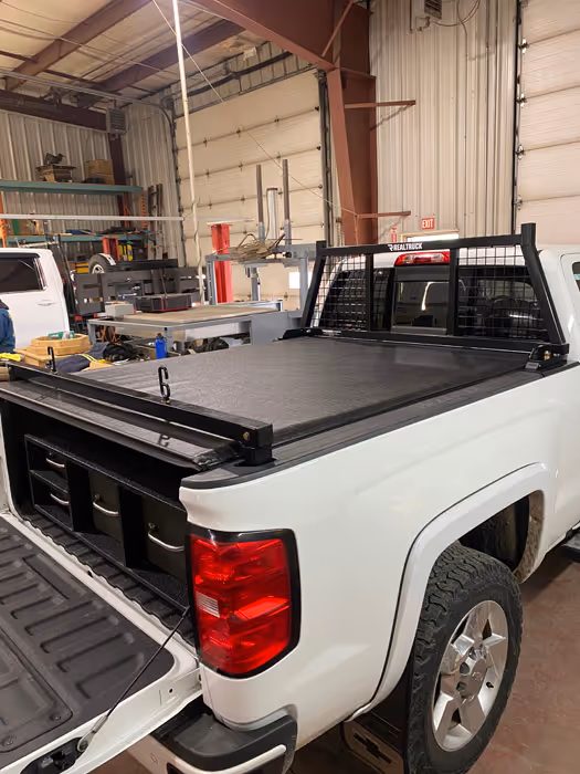 White pickup truck with an open tailgate, equipped with a black headache rack, back bar, and a closed tonneau cover on the bed, inside a workshop.