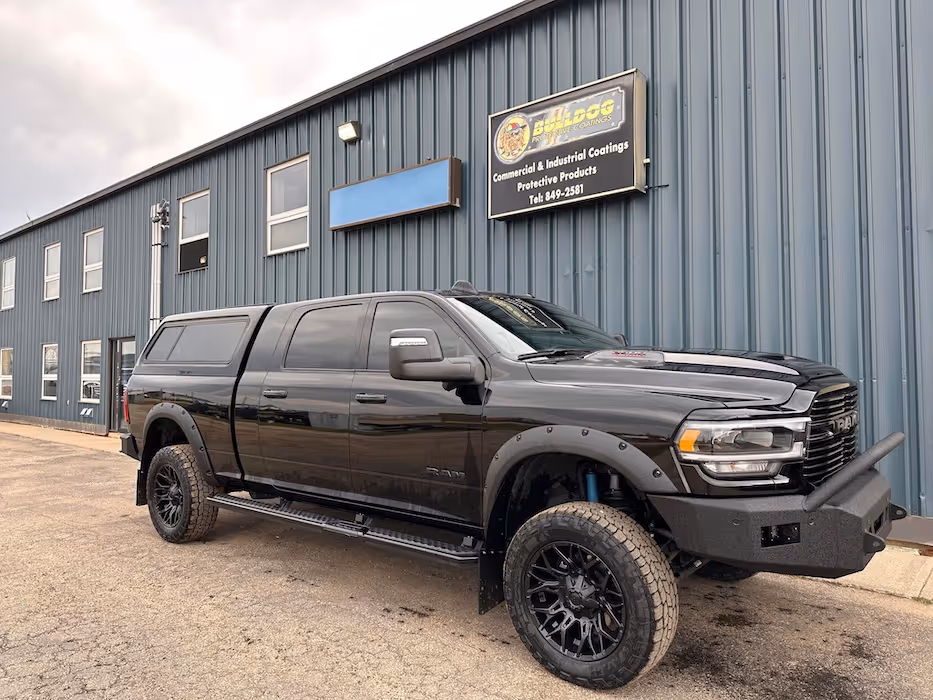 Black RAM pickup truck with off-road tires parked in front of a blue industrial building with a Bulldog Protective Coatings sign.