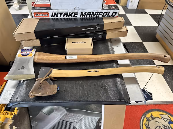 Two axes with wooden handles laid on a black surface; one has a leather sheath on the blade, with boxes and an intake manifold product behind them on a checkered floor.