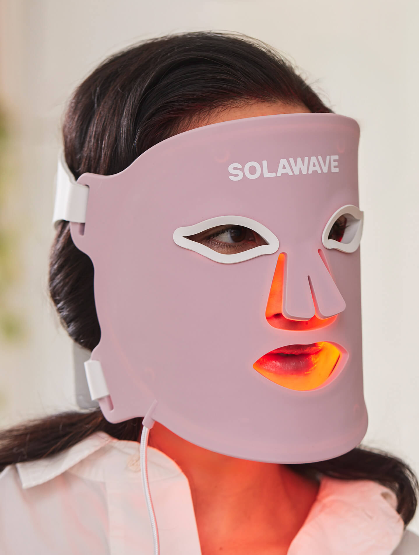 Person wearing a pink LED light therapy facial mask labeled SOLAWAVE with orange light glowing around the nose and mouth openings.