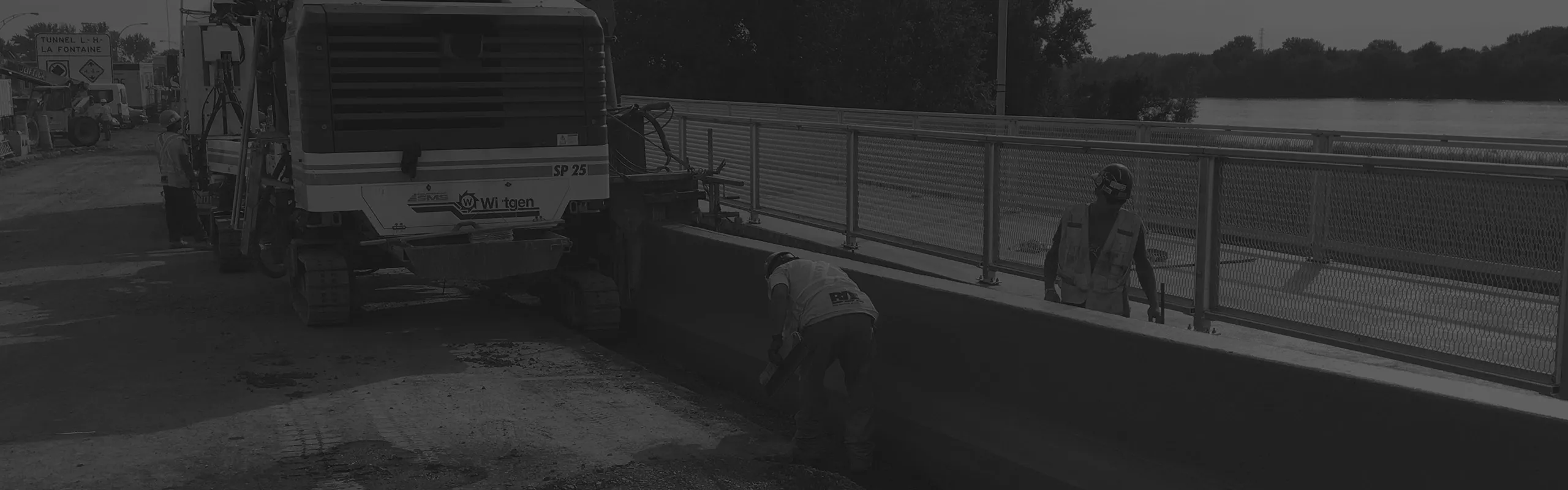 Travailleurs de construction portant un casque et un gilet réfléchissant sur un chantier près d'une barrière métallique à côté d'un cours d'eau.