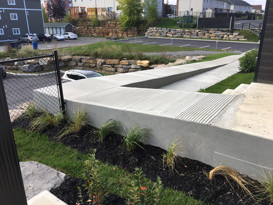 Rampe d'accessibilité en béton longeant un trottoir près d'un stationnement et de maisons résidentielles.