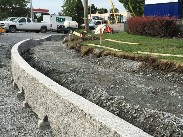 Construction site with a curved stone retaining wall being built along a gravel pathway near grass and parked trucks.