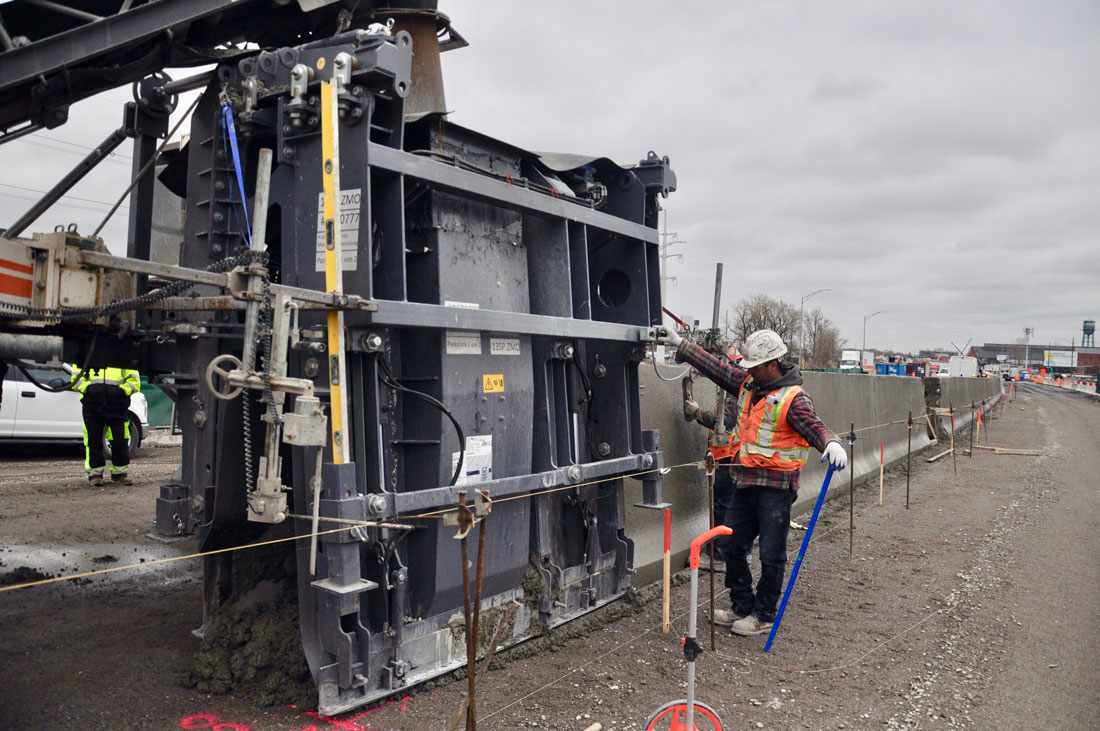 Ouvrier en gilet de sécurité et casque blanc utilisant une grande machine sur un chantier routier sous un ciel nuageux.