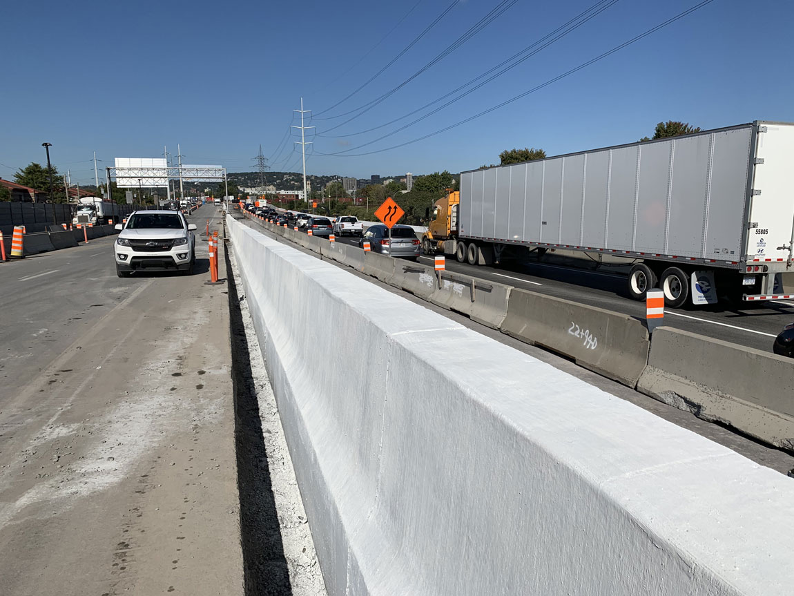 Voie de circulation en construction avec une file de véhicules et un camion-remorque sur l’autoroute, séparés par un mur de béton neuf.