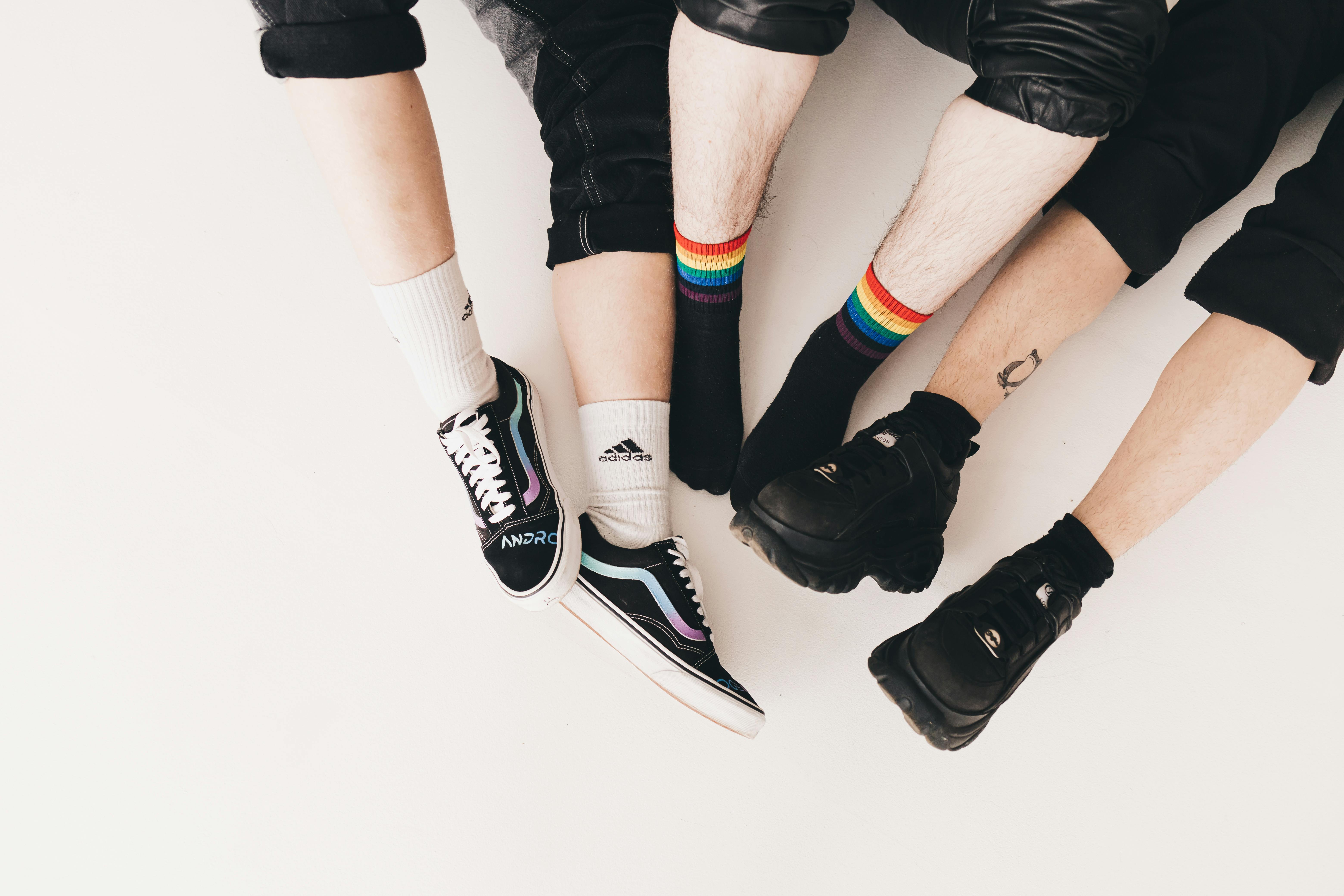 The legs and feet of three people, they all have on black jeans raised to their shins. The person on the left has black shoes with rainbow accents and white socks. The person in the middle has black shoes with rainbow socks. The person on the right has black shoes and a minimal tattoo on their right ankle.