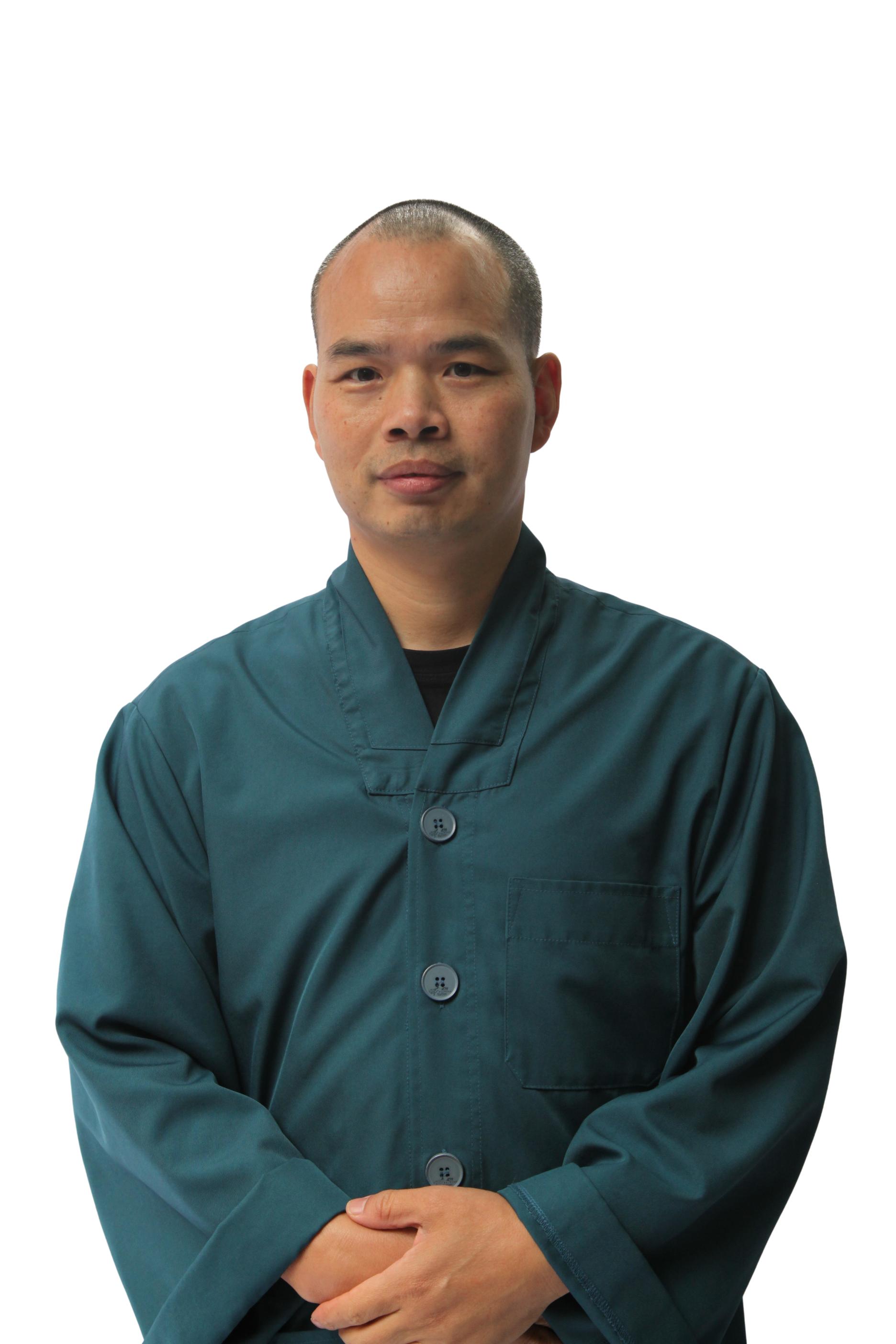 Man with short hair wearing a dark teal buttoned jacket, standing with hands clasped.
