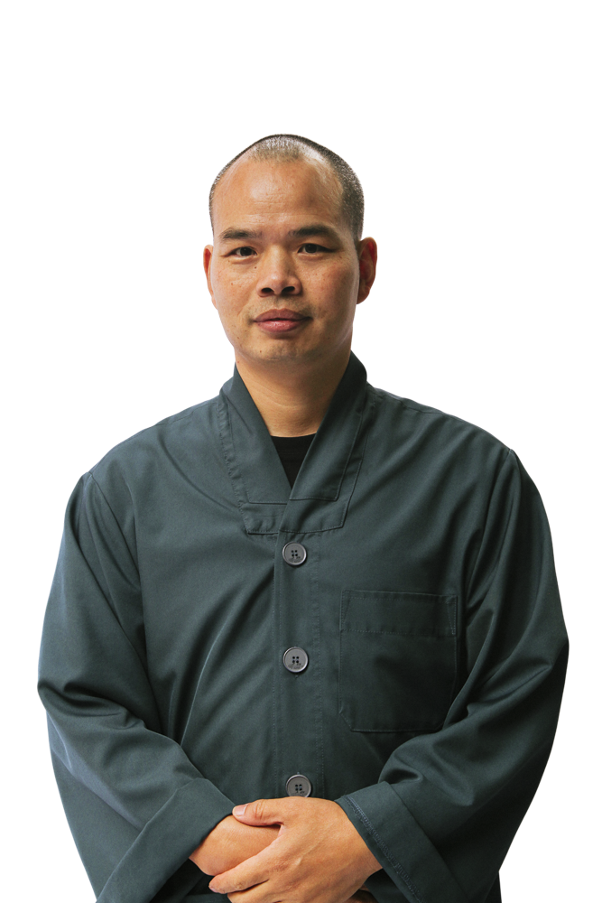 Man with closely shaved hair wearing a dark green buttoned jacket, standing with hands clasped.