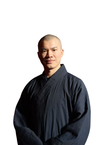 Man with shaved head wearing a dark traditional robe, standing with hands folded.