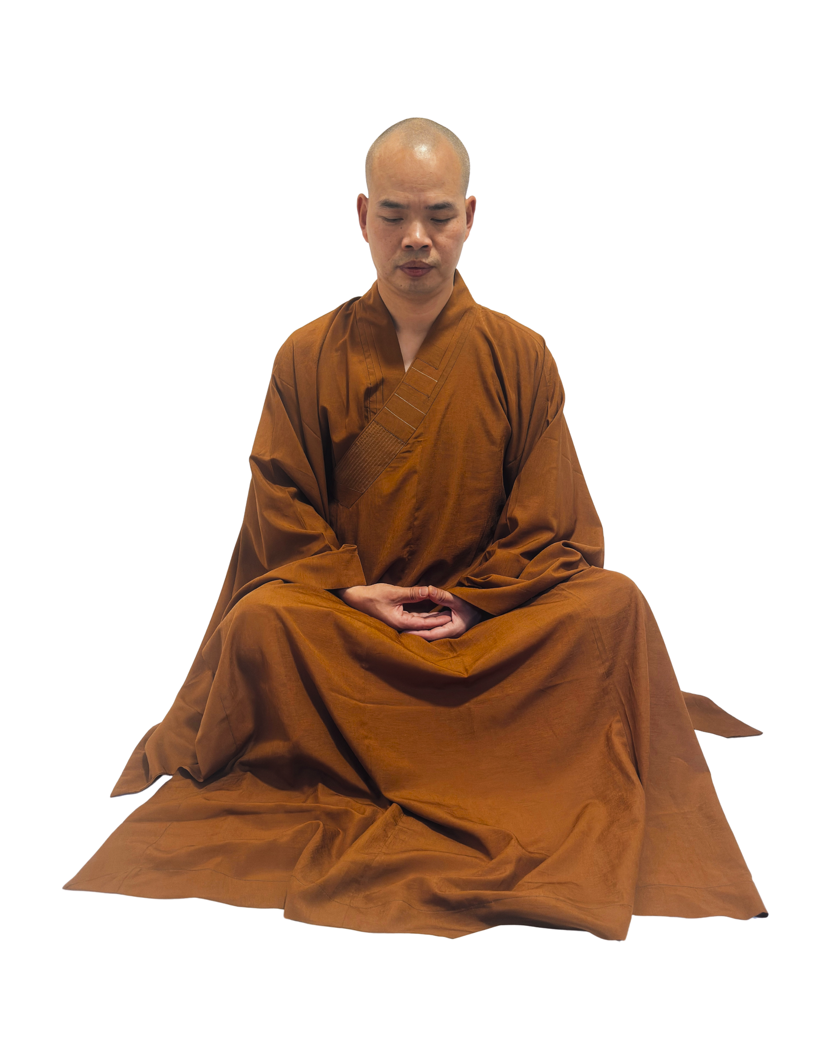 A man with a shaved head wearing a brown robe sits cross-legged with hands in his lap, meditating.