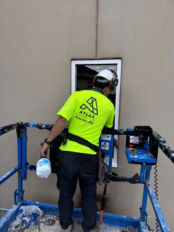 Worker in a neon yellow shirt and white helmet installing or repairing a window while standing on a blue scaffold platform.
