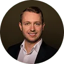 Smiling man with short hair wearing a dark suit jacket and white shirt against a dark background.