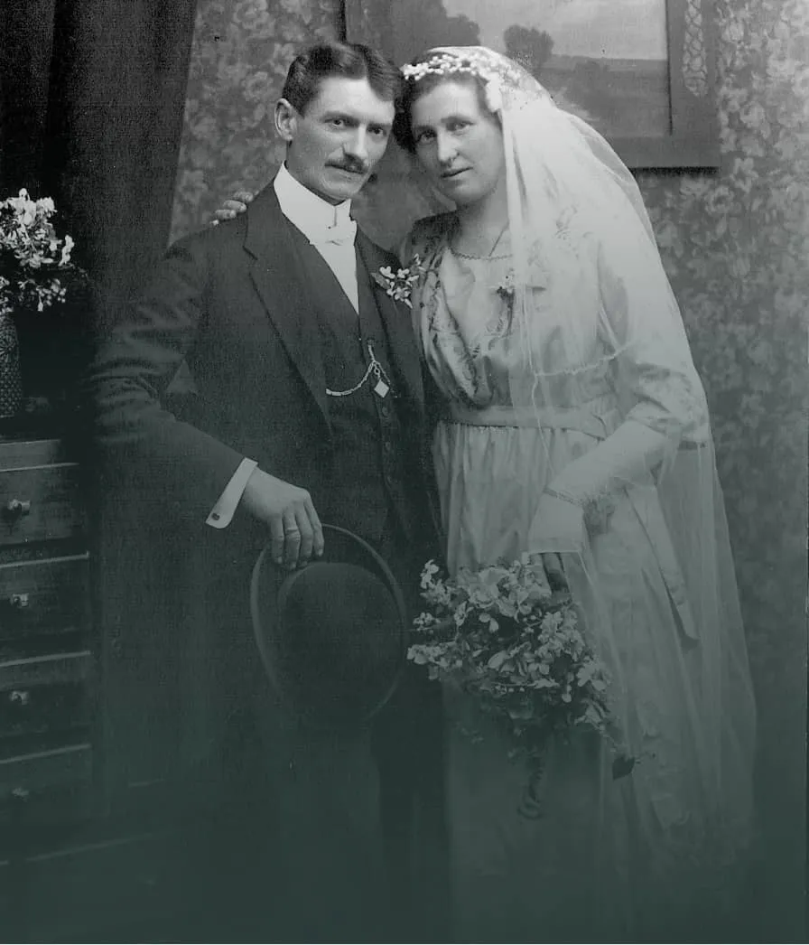 Vintage black-and-white photo of a bride and groom posing indoors, the bride holding a bouquet and wearing a veil, and the groom holding a hat.