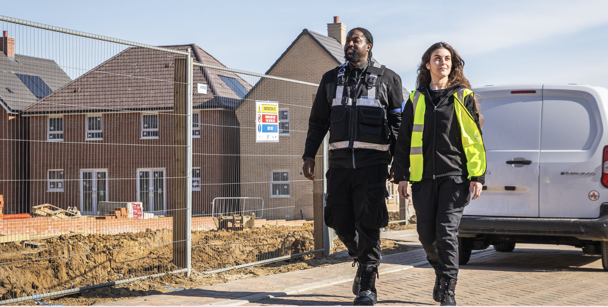 Two AURA security escorts in hi-vis walking the perimeter of a UK construction site of new-build homes, illustrating scheduled security escorting for lone workers