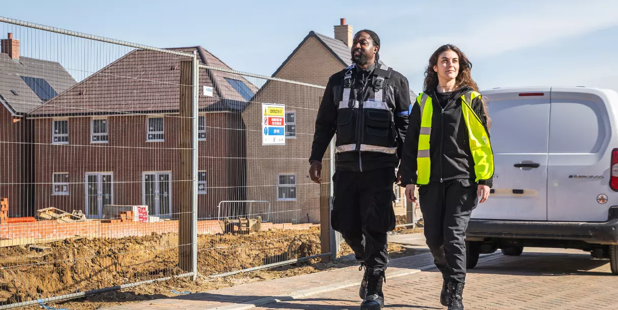 Two AURA security escorts in hi-vis walking the perimeter of a UK construction site of new-build homes, illustrating scheduled security escorting for lone workers.