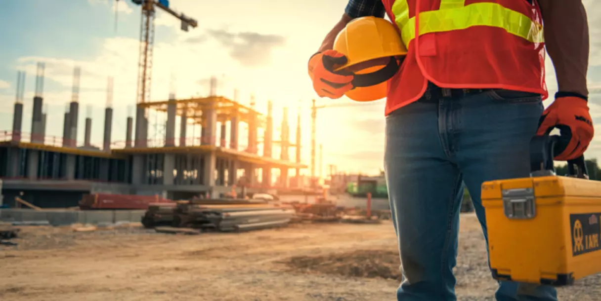 Construction worker holding a hard hat and toolbox at a building site at sunset, representing a lone worker arriving at an unfamiliar or isolated site.