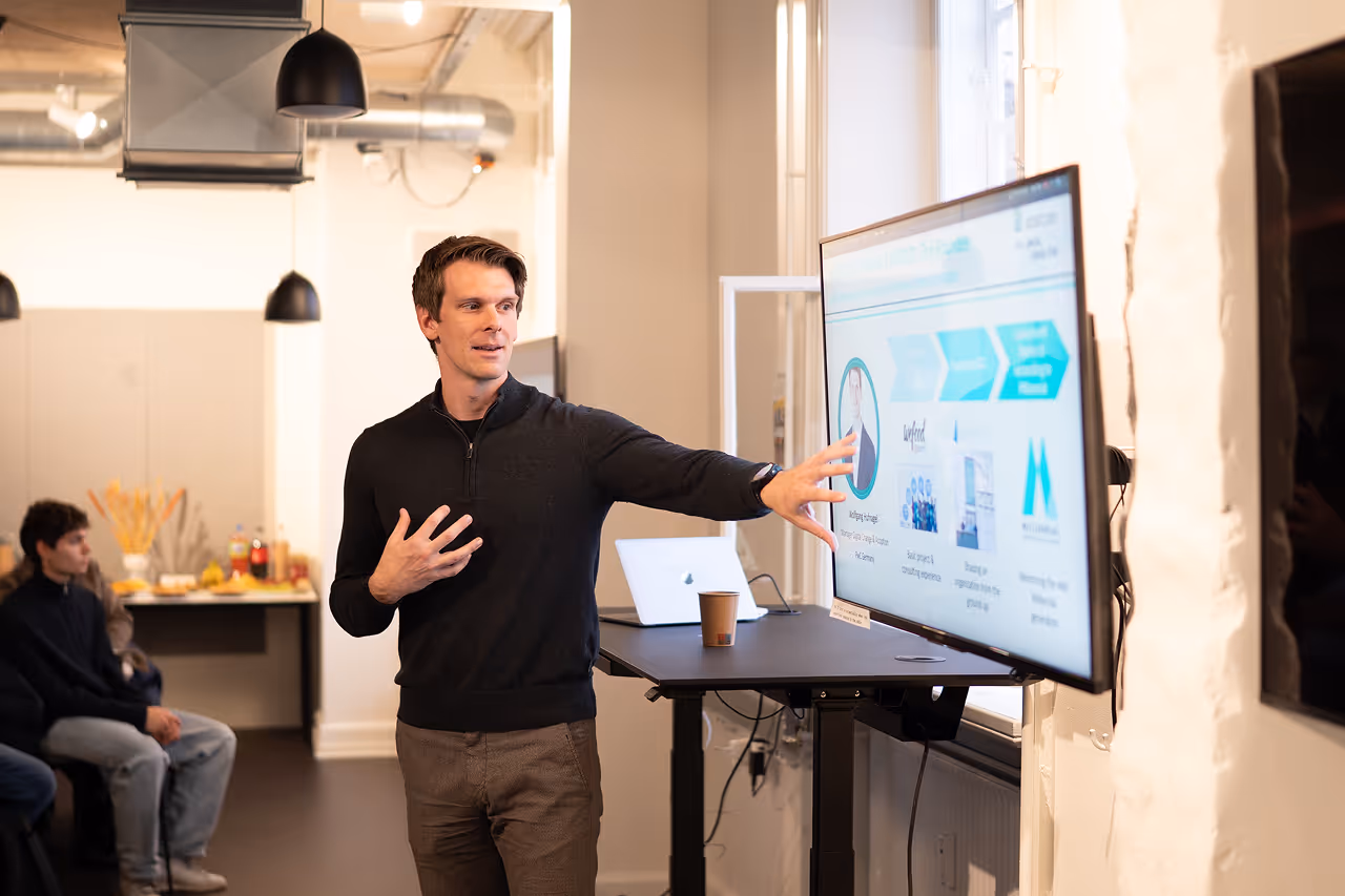 Man giving a presentation pointing at a large screen in a modern office room with seated audience.