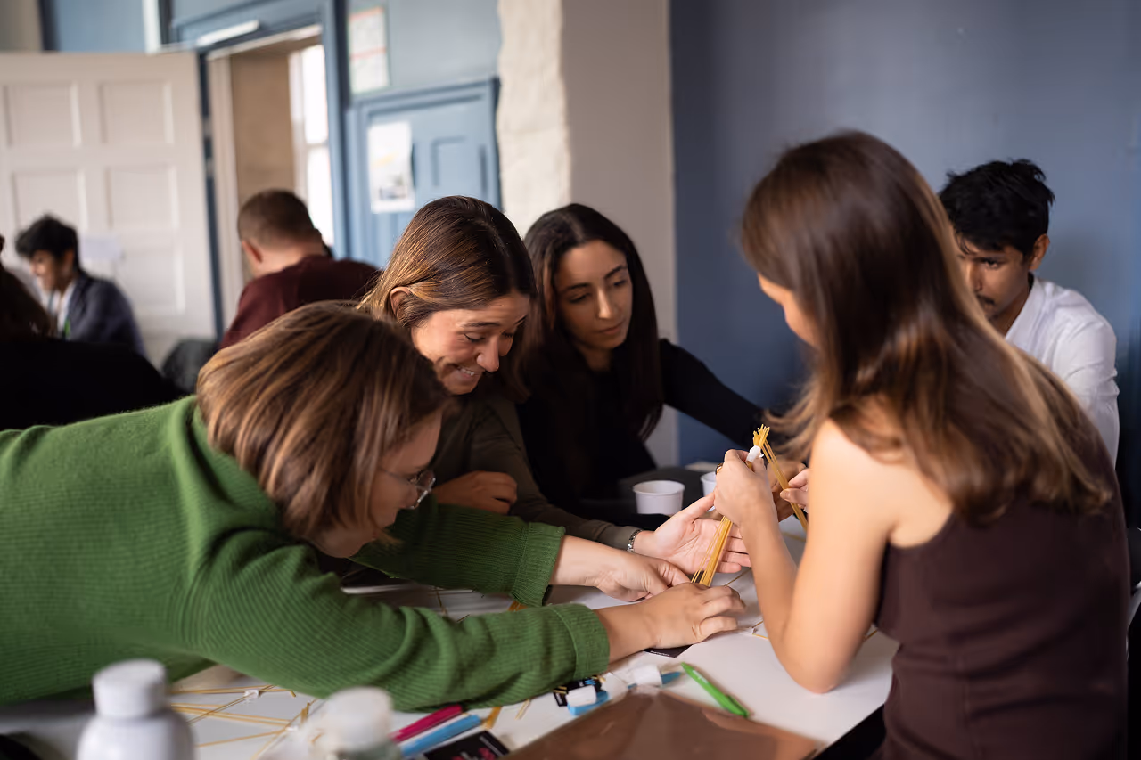 A group of adults collaborating on a hands-on project using dry spaghetti noodles on a table indoors.