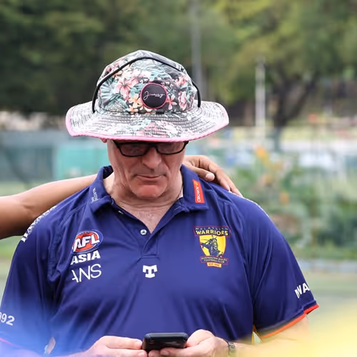 Man wearing a floral bucket hat and blue Warriors sports shirt looking down at his phone, with a hand resting on his shoulder.