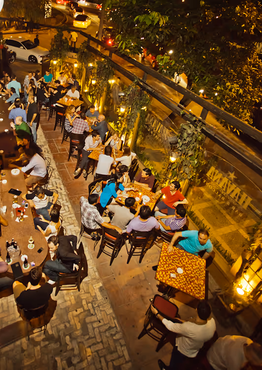 Outdoor evening scene of Havana, people dining at tables alongside a busy street with warm yellow lighting and greenery.