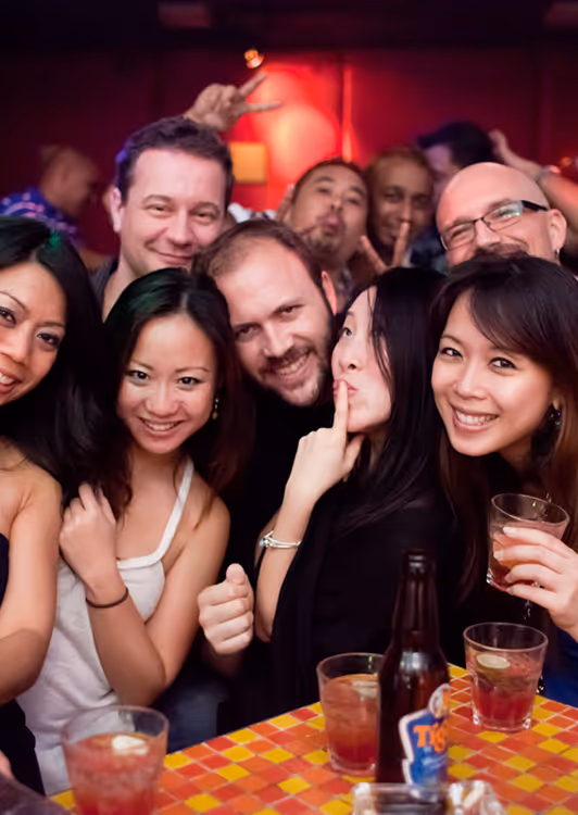 Group of friends smiling and posing closely together at a table with drinks in a lively indoor setting.