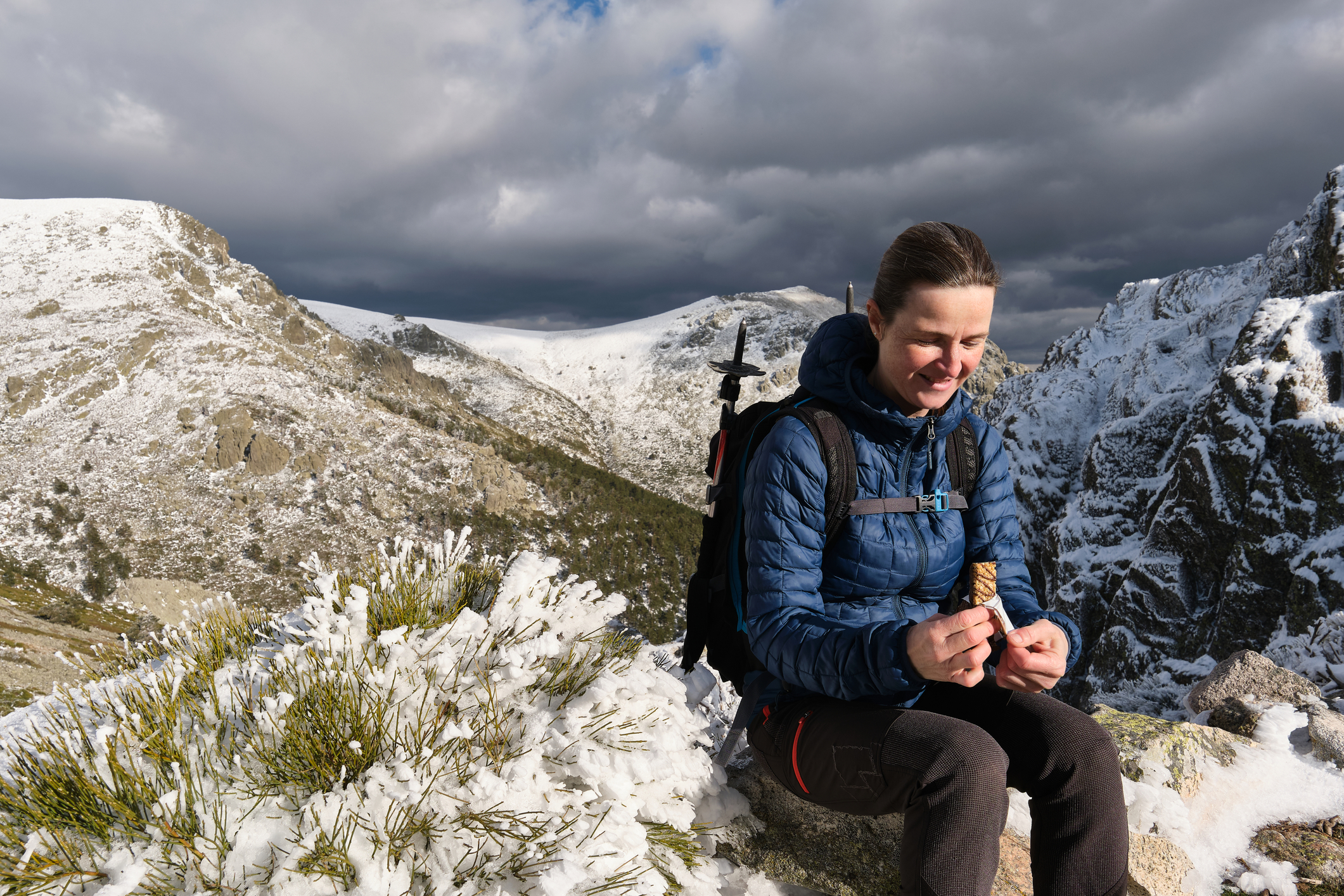 Ours. Glad female backpacker/hiker eating snack while sitting on snowy mountain