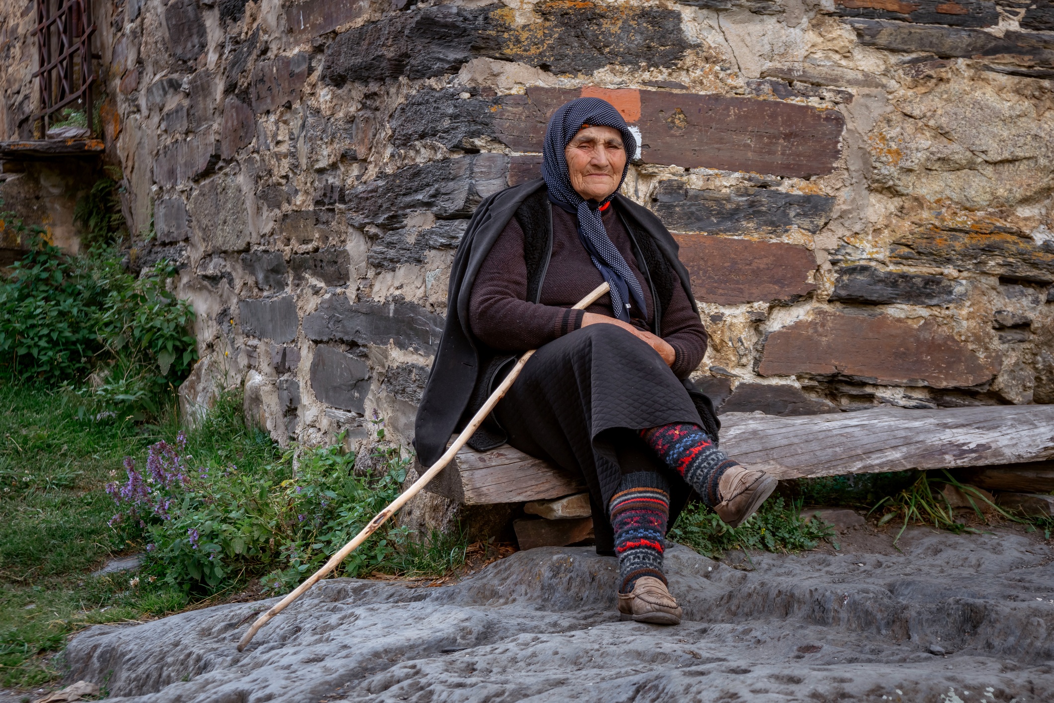 Elderly woman sits in front of the Svan tower with a wooden cane