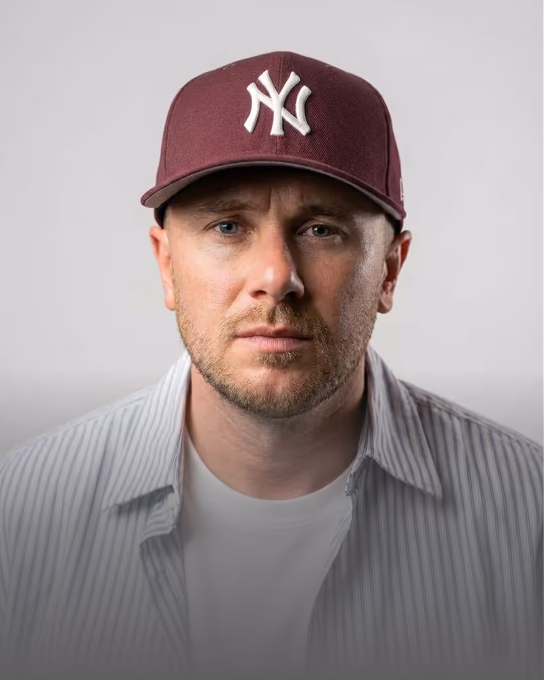 Man with light beard wearing a maroon New York Yankees cap and a striped shirt over a white t-shirt, facing forward against a plain background.