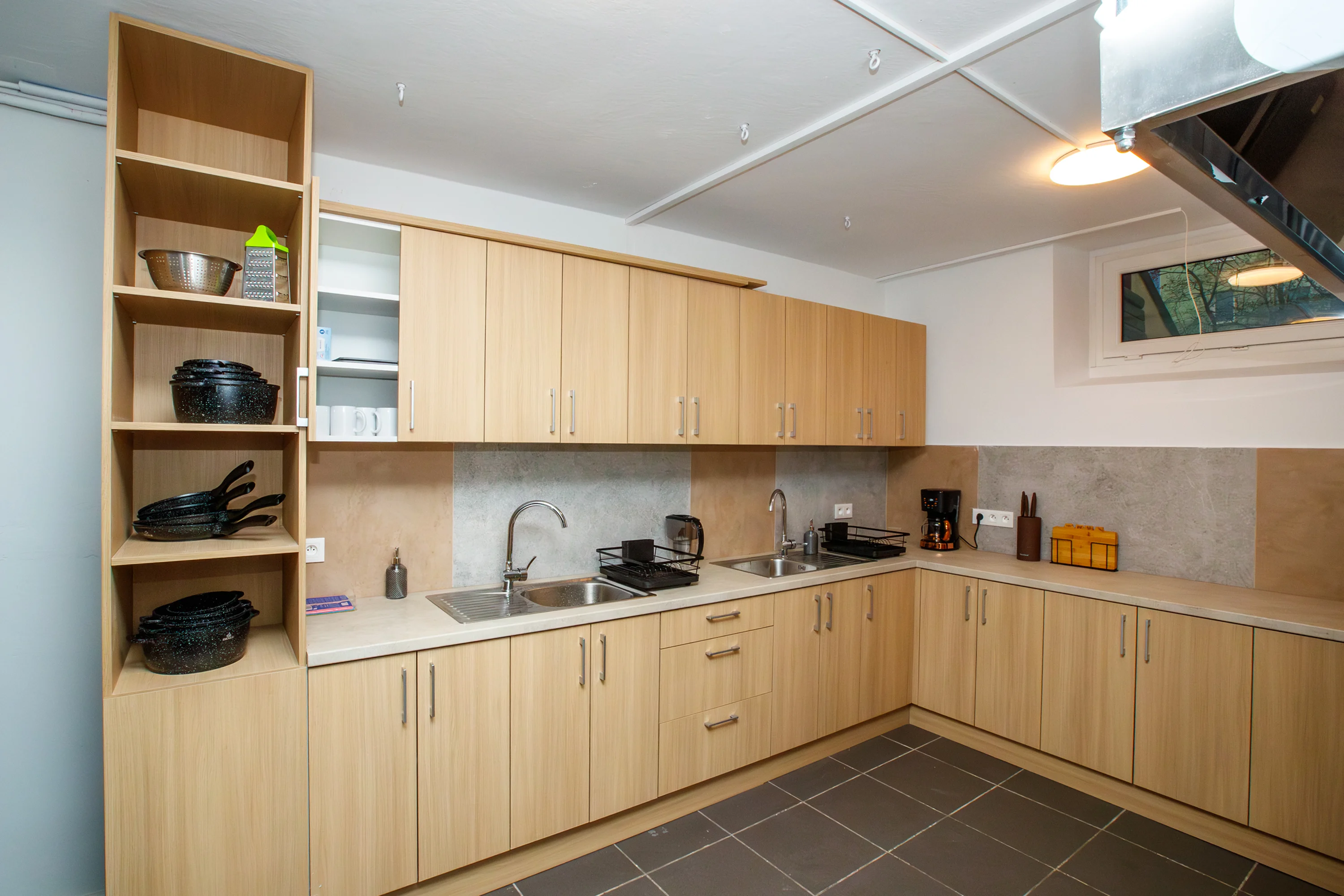 Modern kitchen with light wood cabinets, double sinks, shelves with cookware, coffee maker, and a window above the countertop.