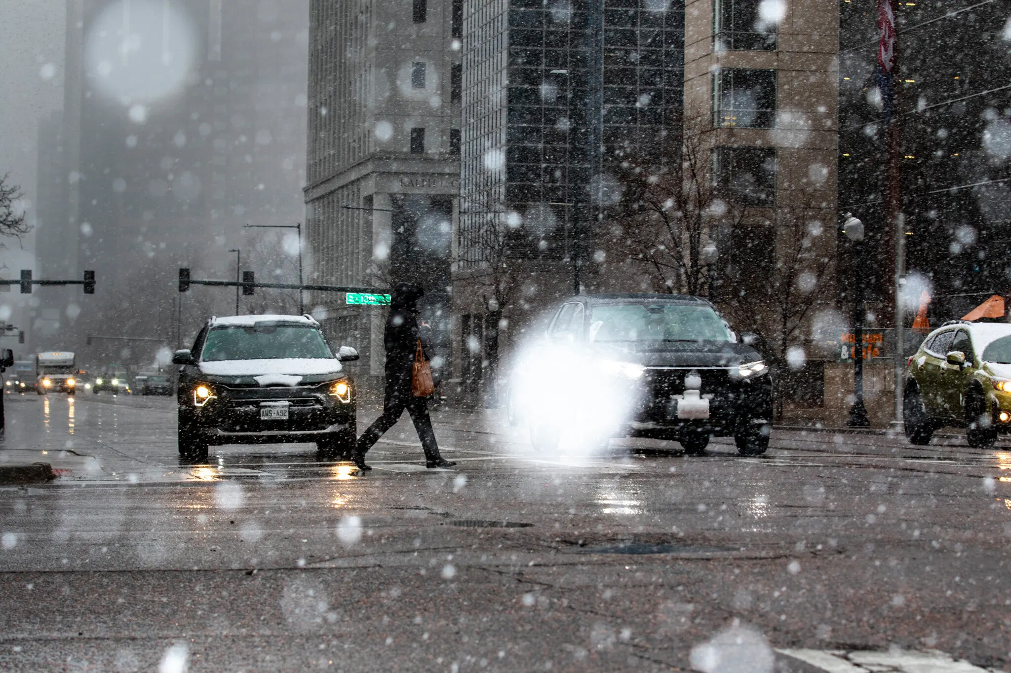 Pedestrian crossing a snowy downtown Denver intersection between two vehicles, one with its high beams on