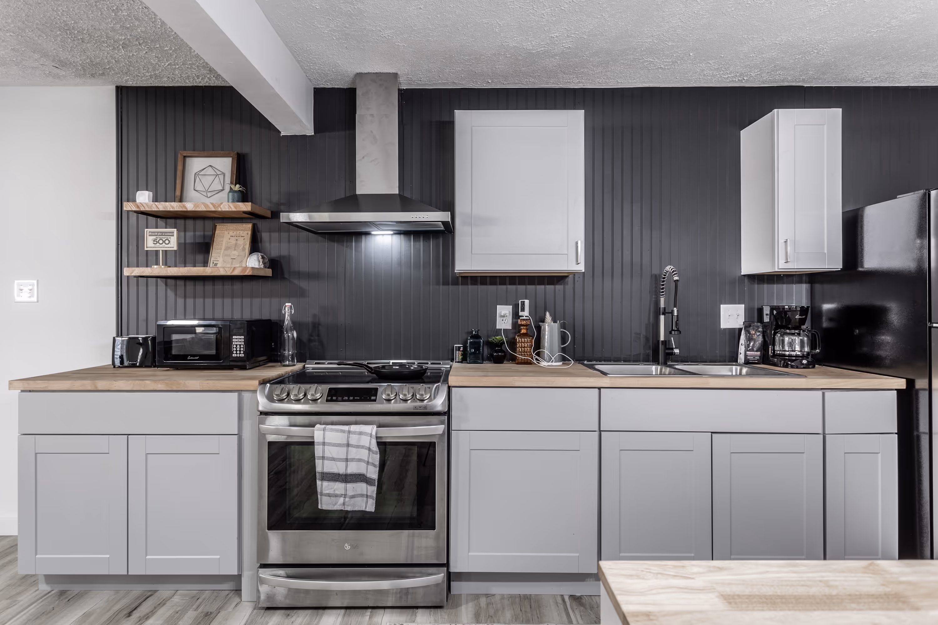 Modern kitchen with stainless steel stove, gray cabinets, wooden countertops, and black backsplash with shelves and appliances.