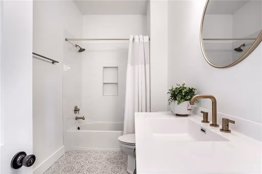 Minimalist bathroom with white walls, patterned tile floor, white bathtub with shower, white curtain, and sink with bronze fixtures and a round mirror.
