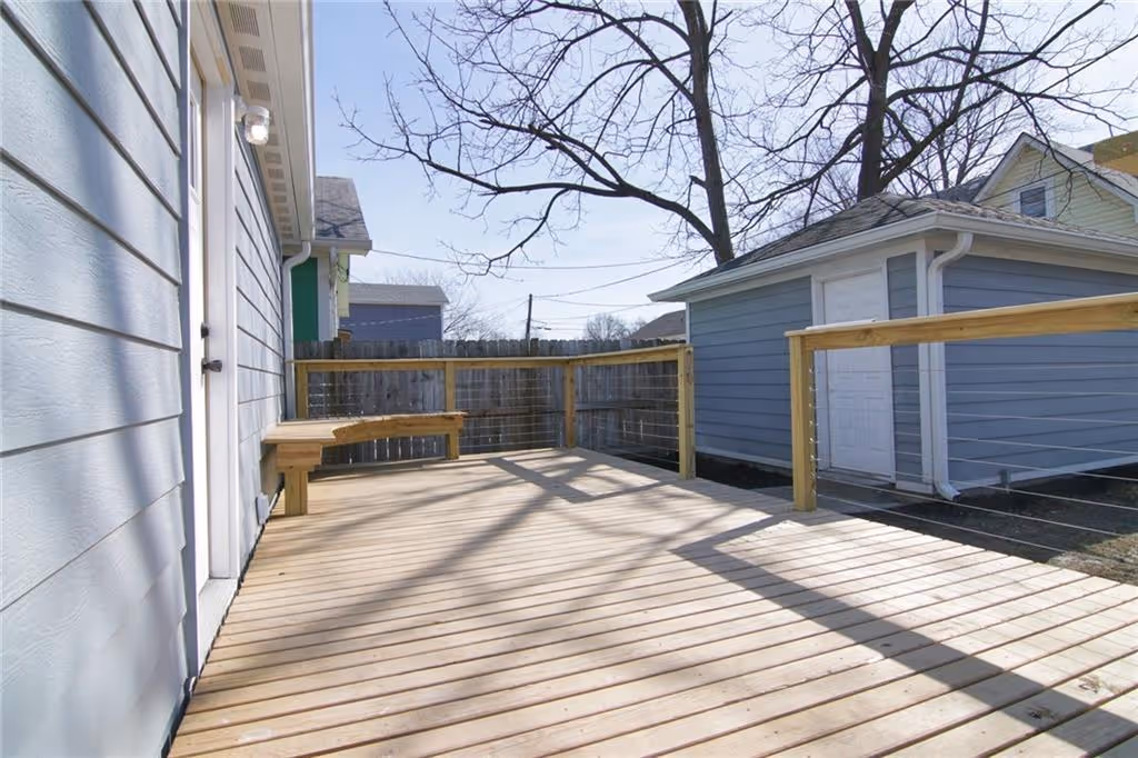Sunny wooden backyard deck attached to a house with a built-in bench and cable railing, next to a detached garage.